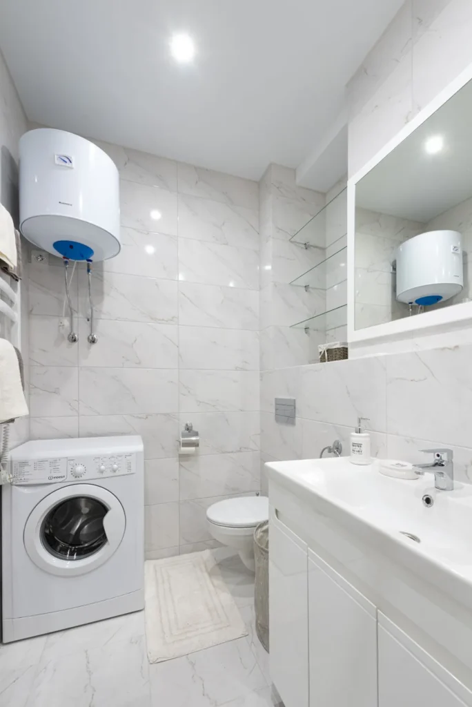 White electric tank water heater installed above a washing machine in a clean bathroom